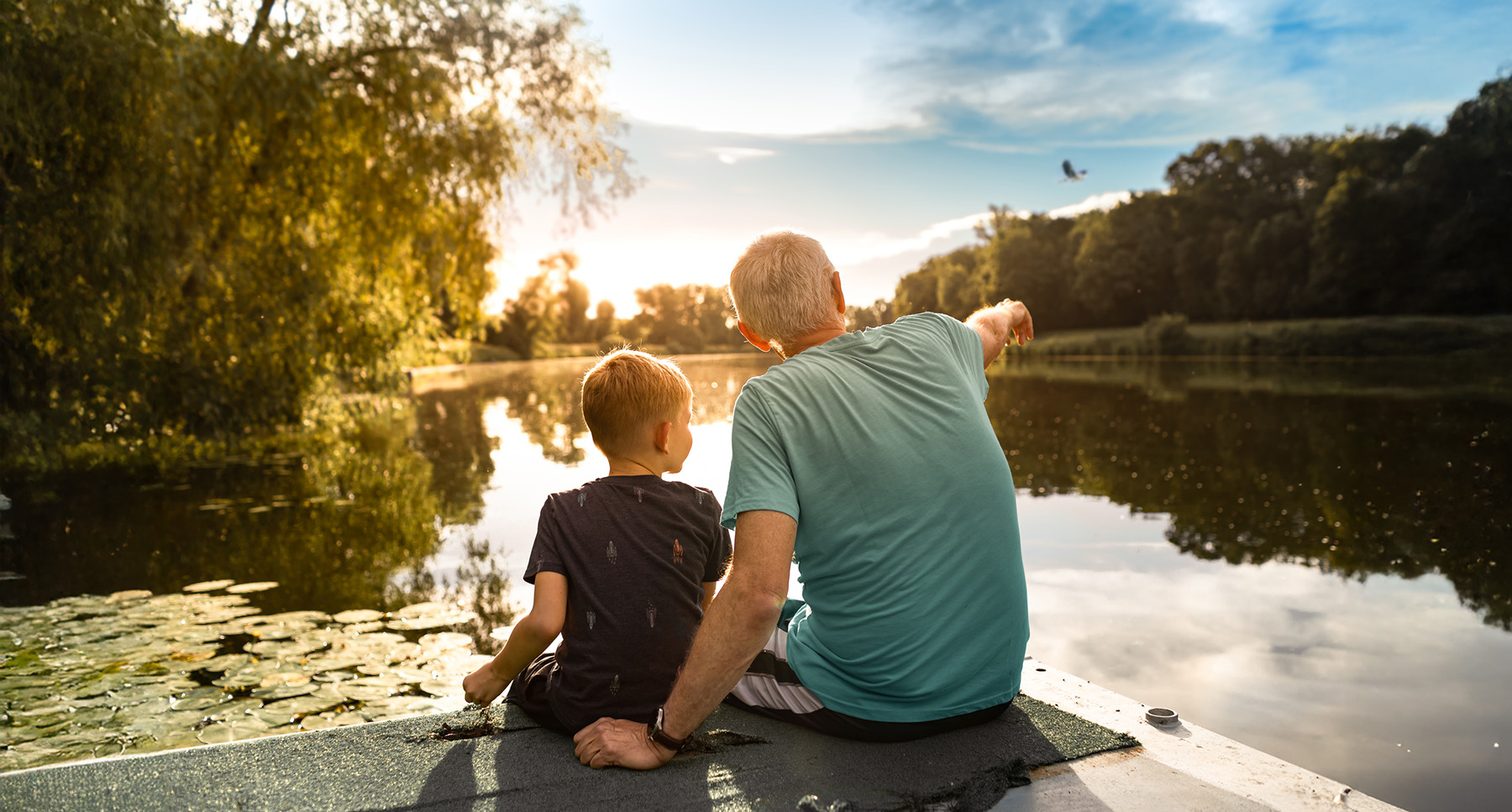 Grandfather and young boy sitting together on a dock, looking out over a lake surrounded by trees at sunset.